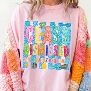 May include: A light pink t-shirt featuring a graphic with blue vertical stripes and the text "CLASS DISMISSED HAVE AN AMAZING SUMMER" in colorful, scribbled-style letters. The design includes a globe, stars, flowers, a smiley face, and pencils.