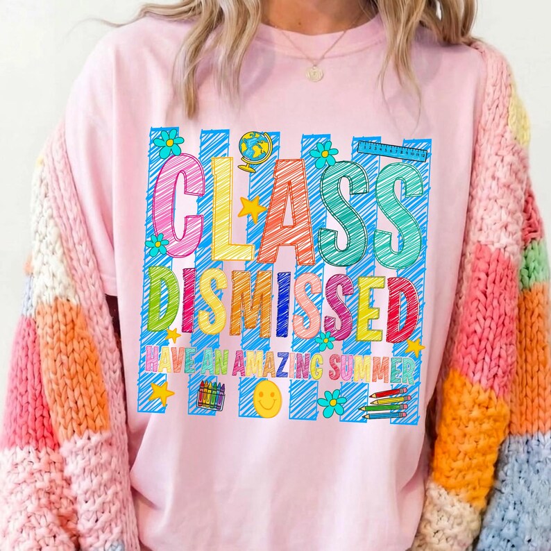 May include: A light pink t-shirt featuring a graphic with blue vertical stripes and the text "CLASS DISMISSED HAVE AN AMAZING SUMMER" in colorful, scribbled-style letters. The design includes a globe, stars, flowers, a smiley face, and pencils.