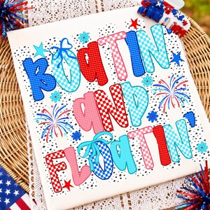 Op de afbeelding: Een wit t-shirt met de tekst "BOATIN AND FLOATIN" in een speels, meerkleurig lettertype met rode, blauwe en roze ruitjes- en stippenpatronen. Het ontwerp bevat kleine blauwe bloemetjes, rode sterren en afbeeldingen van vuurwerk.