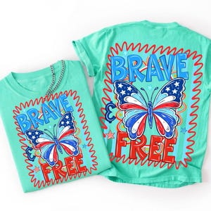 May include: Two mint green t-shirts featuring a graphic design with the text "BRAVE & FREE" in blue and red. The design includes a butterfly with wings patterned in red, white, and blue stars and stripes, surrounded by a red squiggly border.