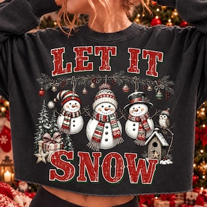 Pu&ograve; includere: Una felpa grigio antracite con la grafica di tre pupazzi di neve che indossano sciarpe e cappelli, con la scritta LET IT SNOW in rosso. Il design include un albero di Natale, un pacco regalo, una casetta per uccelli e un gufo con decorazioni.