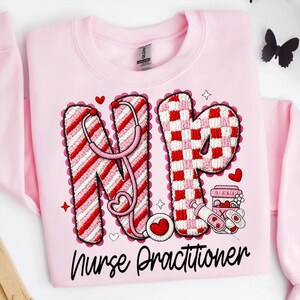 May include: Pink sweatshirt with a graphic design featuring the letters "NP" in red and white, a stethoscope, hearts, and the words "Nurse Practitioner". The design is surrounded by a scalloped border.