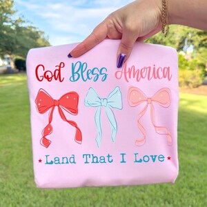 May include: A light pink t-shirt featuring the red and blue text God Bless America above three illustrated bows in red, light blue, and soft pink. Below the bows, the text Land That I Love is printed in blue, flanked by two small red stars.
