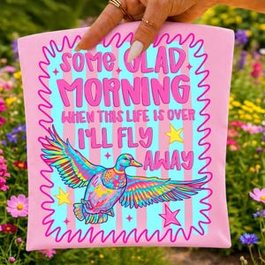 Pu&ograve; includere: Una borsa tote rosa chiaro con sfondo azzurro e strisce verticali rosa. Il testo SOME GLAD MORNING WHEN THIS LIFE IS OVER I'LL FLY AWAY &egrave; stampato in rosa. Sono presenti un'anatra colorata con ali spiegate e delle stelle gialle.