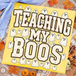 May include: A square yellow sign featuring the text TEACHING MY BOOS in large black-outlined block letters with a speckled texture. The design is surrounded by numerous small white ghost illustrations with black eyes and mouths on a yellow background.