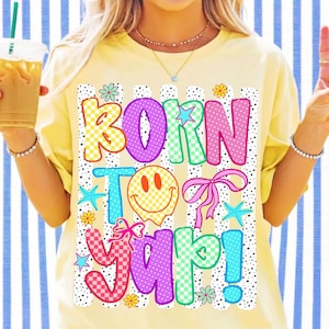 May include: A pale yellow t-shirt featuring a graphic print with the text BORN TO YAP! in colorful, patterned block letters. The design includes a yellow checkered smiley face, pink bows, small blue starfish, and flowers over a dotted background.