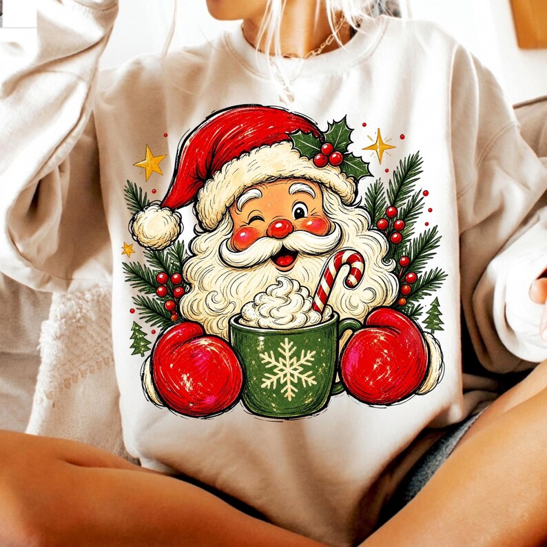 May include: A cream-coloured crewneck sweatshirt featuring a graphic of Santa Claus wearing red mittens and a red hat. Santa is winking and holding a green mug with a white snowflake design, filled with whipped cream and a candy cane, surrounded by pine.