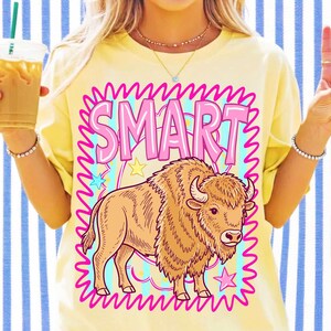 May include: A pale yellow t-shirt featuring a graphic print of a brown bison. Above the bison, the word SMART is written in large pink letters. The design is enclosed in a bright pink squiggly border with small yellow and blue stars.