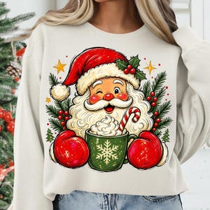 May include: A cream-coloured crewneck sweatshirt featuring a graphic print of Santa Claus wearing a red hat and red mittens. Santa is winking and holding a green mug filled with whipped cream and a candy cane, surrounded by pine branches and stars.