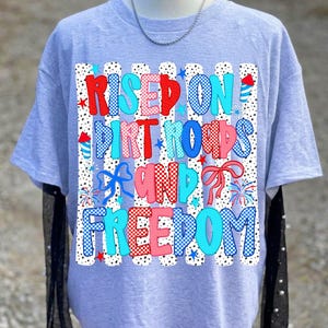 May include: A light grey t-shirt featuring a graphic print with the text "RISED ON DIRT ROADS AND FREEDOM". The design includes red, white, and blue patterns, polka dots, stripes, stars, fireworks, and bows, arranged in a patriotic style.