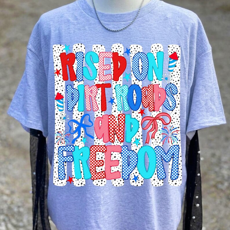 May include: A light grey t-shirt featuring a graphic print with the text "RISED ON DIRT ROADS AND FREEDOM". The design includes red, white, and blue patterns, polka dots, stripes, stars, fireworks, and bows, arranged in a patriotic style.