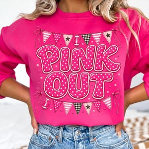 K&ouml;nnte beinhalten: Ein leuchtend pinkfarbenes Langarm-Sweatshirt mit dem Schriftzug PINK OUT in gro&szlig;en, wei&szlig; gepunkteten pinken Buchstaben. Das Design enth&auml;lt eine dekorative Wimpelkette mit Fu&szlig;b&auml;llen, Sternen und Karomustern in Pink, Wei&szlig; und Schwarz.