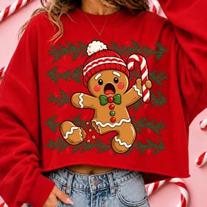 May include: A red long-sleeve sweatshirt featuring a graphic of a gingerbread person wearing a red and white knit hat, holding a candy cane. The gingerbread person has a missing leg and is set against a background of green pine branches.