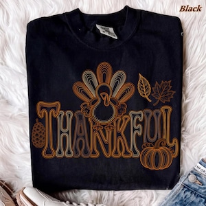 May include: A black short-sleeved t-shirt featuring an orange line-art graphic of a turkey, a pinecone, autumn leaves, and a pumpkin. The word THANKFUL is printed in a stylized orange font integrated with the turkey illustration.