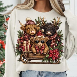 Puede incluir: Una sudadera de color crema con un gr&aacute;fico de tres vacas Highland en un trineo de madera. Las vacas llevan gorros de invierno, bufandas y gafas de sol, rodeadas de ramas de pino, bayas rojas, cajas de regalo y una gran flor de pascua roja.