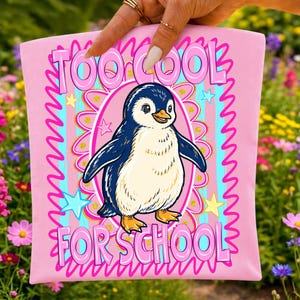 Pu&ograve; includere: Una pochette in tessuto rosa chiaro con un'illustrazione centrale di un pinguino all'interno di una cornice ovale rosa. Il testo TOO COOL FOR SCHOOL &egrave; stampato in grandi lettere rosa sopra e sotto il pinguino. Lo sfondo ha strisce blu e stelle.