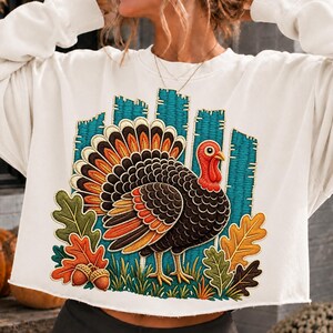 May include: A cream-colored long-sleeve sweatshirt featuring a large embroidered graphic of a turkey with brown, orange, and yellow feathers. The design includes a teal fence background, green grass, and colorful autumn oak leaves with two acorns.