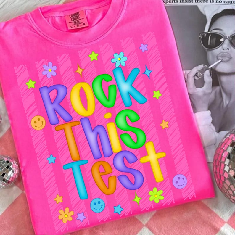 May include: Bright pink t-shirt with the phrase "ROCK THIS TEST" in colorful bubble letters. The shirt features a vertical striped pattern and is decorated with stars, flowers, and smiley faces. The shirt is a Comfort Colors brand.