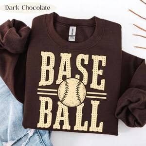 Puede incluir: Una sudadera de cuello redondo en color marr&oacute;n chocolate oscuro con el texto BASE BALL en una fuente grande de color crema con estilo perforado. Un gr&aacute;fico de una pelota de b&eacute;isbol se sit&uacute;a en el centro entre las palabras, sustituyendo la A.