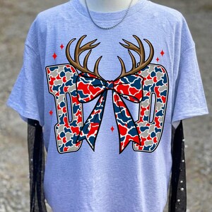 May include: A light grey short-sleeve t-shirt featuring a large graphic of the word DAD in a red, white, and blue camouflage pattern. The letters are accented with brown deer antlers and a matching camouflage bow, surrounded by small red stars.