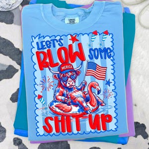 May include: A light blue t-shirt featuring a graphic of a highland cow wearing a red, white, and blue hat and heart-shaped sunglasses, sitting in a pool float. The text reads "LET'S BLOW SOME SHIT UP" with a flag, fireworks, and bottle illustrations.