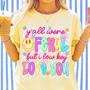 May include: A pale yellow short-sleeve t-shirt featuring a graphic print with black vertical stripes and dots. The text reads y'all were FERAL but i low key LOVE YOU, accompanied by a yellow smiley face, small stars, flowers, and bows.