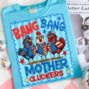 May include: A light blue t-shirt featuring a graphic of three chickens wearing patriotic American flag-themed hats, sunglasses, and bandanas. The text reads BANG BANG MOTHER CLUCKERS in red and blue lettering, surrounded by stars and a scalloped border.