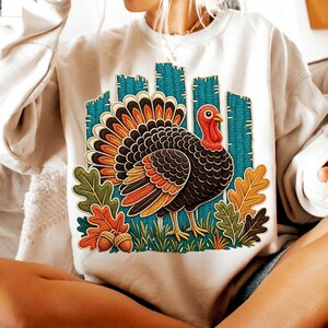 May include: A cream-colored crewneck sweatshirt featuring a central graphic of a turkey with black, orange, and brown feathers. The turkey stands amidst green grass, colorful autumn leaves, and two acorns, set against a teal vertical striped background.
