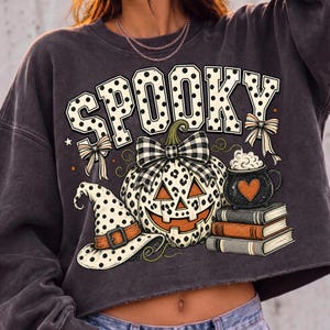 May include: A charcoal gray long-sleeve sweatshirt featuring a graphic print with the word SPOOKY in polka-dot letters. The design includes a leopard-print pumpkin with a plaid bow, a polka-dot witch hat, a stack of books, and a cauldron.