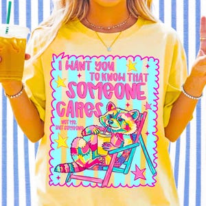 K&ouml;nnte beinhalten: Ein blassgelbes T-Shirt mit einer bunten Grafik eines Waschb&auml;ren, der in einem Liegestuhl sitzt und ein Getr&auml;nk h&auml;lt. Der Text lautet: I WANT YOU TO KNOW THAT SOMEONE CARES NOT ME, BUT SOMEONE. Das Design enth&auml;lt Sterne und einen gewellten Rand.