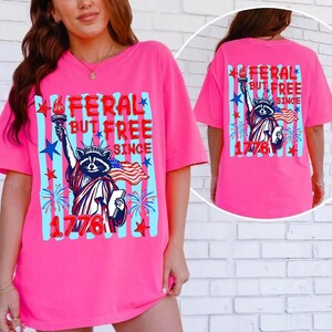 Może przedstawiać: Jaskrawor&oacute;żowy t-shirt z grafiką szopa pracza przebranego za Statuę Wolności, trzymającego pochodnię i książkę. Napis głosi FERAL BUT FREE SINCE 1776, w towarzystwie niebieskich i czerwonych gwiazd, fajerwerk&oacute;w oraz powiewającej amerykańskiej flagi.