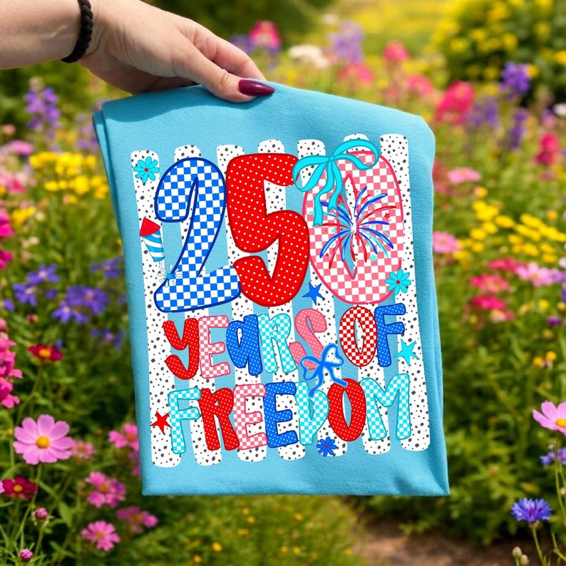May include: A light blue t-shirt featuring a graphic design with the text 250 YEARS OF FREEDOM. The design includes red, white, and blue checkered and polka-dot patterns, firework illustrations, and light blue bows on a white striped background.