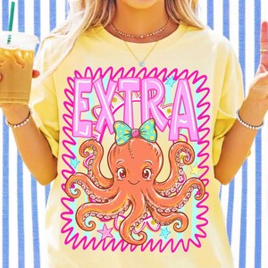 May include: A pale yellow t-shirt featuring a graphic print of an orange octopus wearing a teal and yellow polka-dot bow. The word EXTRA is printed in large pink letters above the octopus, all enclosed within a bright pink squiggly border.