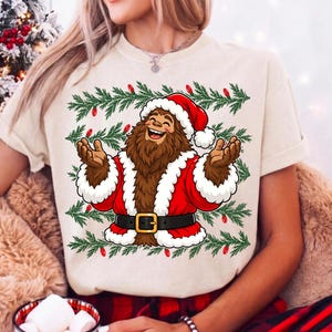 Puede incluir: Una camiseta de manga corta color crema con un gr&aacute;fico de un Bigfoot marr&oacute;n vestido con un traje de Pap&aacute; Noel rojo con ribete de piel blanca y un cintur&oacute;n negro. El dise&ntilde;o est&aacute; rodeado de ramas de pino verdes y peque&ntilde;as luces navide&ntilde;as rojas.