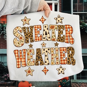 May include: A white t-shirt featuring the text "SWEATER WEATHER" in large, bold letters. Each letter is filled with a variety of patterns including orange plaid, leopard print, polka dots, and floral designs. Small stars and diamond shapes surround the text.