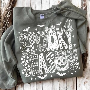 May include: An olive green crewneck sweatshirt featuring a white graphic print that reads SPOOKY SEASON. The design includes a ghost with a bow, a jack-o'-lantern with a bow, a witch hat, bats, and stars, with letters filled with floral, checkered, and striped patterns.