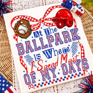 Op de afbeelding: Een wit T-shirt met een rood-blauwe afbeelding van een honkbalhandschoen, een honkbal en een rode slaghelm. De tekst luidt At The BALLPARK Is Where I Spend Most OF MY DAYS, geaccentueerd door rood-blauwe geblokte linten en bliksemschichten.