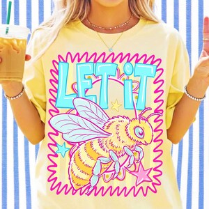 Peut inclure: Un t-shirt jaune p&acirc;le avec une impression graphique repr&eacute;sentant une abeille aux ailes bleu clair et au corps jaune. Le texte "LET IT" est imprim&eacute; en grandes lettres bleu clair au-dessus de l'abeille. Le motif est entour&eacute; d'une bordure rose.