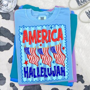 Puede incluir: Una camiseta de cuello redondo azul claro con un estampado gr&aacute;fico que muestra el texto rojo AMERICA sobre tres ilustraciones de banderas estadounidenses colgadas en un tendedero, con el texto azul HALLELUJAH debajo, enmarcado por un borde festoneado.