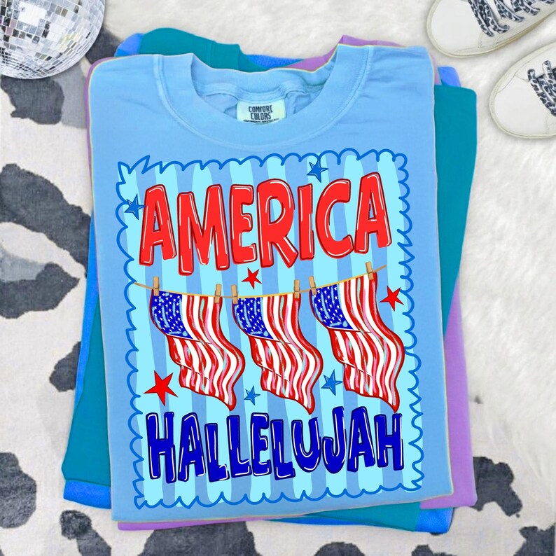 Puede incluir: Una camiseta de cuello redondo azul claro con un estampado gr&aacute;fico que muestra el texto rojo AMERICA sobre tres ilustraciones de banderas estadounidenses colgadas en un tendedero, con el texto azul HALLELUJAH debajo, enmarcado por un borde festoneado.