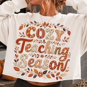 May include: A white long-sleeve sweatshirt featuring the text "Cozy Teaching Season" in large, decorative letters. The lettering incorporates orange, brown, and cream patterns, surrounded by a detailed border of autumn leaves and flowers.