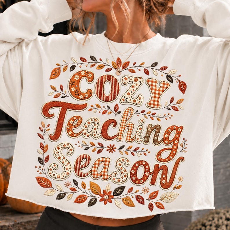 May include: A white long-sleeve sweatshirt featuring the text "Cozy Teaching Season" in large, decorative letters. The lettering incorporates orange, brown, and cream patterns, surrounded by a detailed border of autumn leaves and flowers.