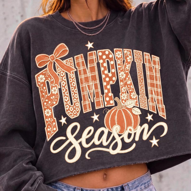 May include: A charcoal gray cropped sweatshirt featuring the text PUMPKIN SEASON in a decorative font. The word PUMPKIN is filled with orange and white patterns including plaid, polka dots, and flowers, accented by a bow and a pumpkin graphic.