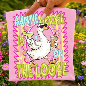 K&ouml;nnte beinhalten: Rosafarbenes T-Shirt mit einer Cartoon-Gans, die eine Sonnenbrille tr&auml;gt und einen Cocktail und ein Getr&auml;nk mit Strohhalm h&auml;lt. Der Text auf dem Shirt lautet "Auntie Goose Is On The Loose" in einer bunten, wellenf&ouml;rmigen Schrift.
