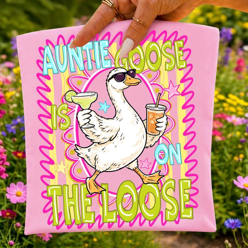 K&ouml;nnte beinhalten: Rosafarbenes T-Shirt mit einer Cartoon-Gans, die eine Sonnenbrille tr&auml;gt und einen Cocktail und ein Getr&auml;nk mit Strohhalm h&auml;lt. Der Text auf dem Shirt lautet "Auntie Goose Is On The Loose" in einer bunten, wellenf&ouml;rmigen Schrift.