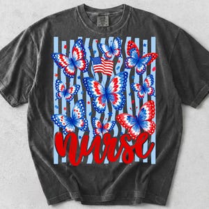 May include: A charcoal grey short-sleeve t-shirt featuring a graphic print of red, white, and blue butterflies, a small American flag, and scattered stars over light blue vertical stripes. The word NURSE is printed in large red script at the bottom.