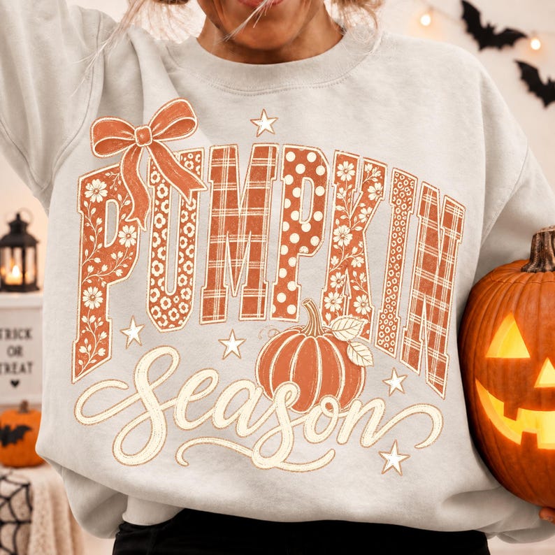 May include: A light gray crewneck sweatshirt featuring the text PUMPKIN SEASON in large block letters with floral, polka dot, and plaid patterns in orange and white. A decorative orange bow sits above the text, and a pumpkin graphic is below.