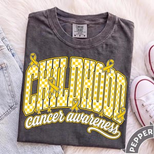 Puede incluir: Una camiseta gris carb&oacute;n con el texto CHILDHOOD en un patr&oacute;n de cuadros amarillos y blancos, con la palabra awareness en letra cursiva blanca debajo. Varios lazos amarillos est&aacute;n integrados en el dise&ntilde;o frontal.