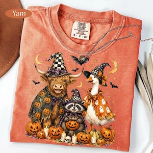 May include: An orange crew neck t-shirt featuring a graphic of a Highland cow, a raccoon, and a goose wearing witch hats and capes. The design includes pumpkins, bats, and crescent moons. The shirt has a Comfort Colors brand label at the neck.