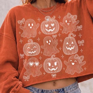 Puede incluir: Una sudadera de manga larga en color naranja quemado con un estampado gr&aacute;fico de l&iacute;neas blancas que muestra varios fantasmas, calabazas y lazos peque&ntilde;os. Los fantasmas tienen diferentes expresiones y las calabazas tienen caras talladas.